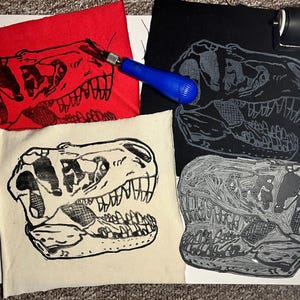 May include: A collection of linocut prints featuring a Tyrannosaurus Rex skull design. The prints are on red, black, and cream-colored fabric. A blue-handled carving tool and a small roller are also visible.