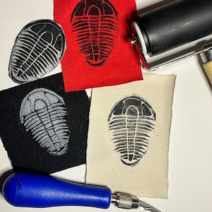 May include: Linocut prints of a trilobite design on red, black, and beige fabric. A black ink roller and a blue-handled carving tool are also present, indicating a printmaking process. The image showcases the art of linocut.