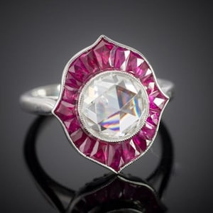 Puede incluir: Un anillo de plata con una gran gema clara y facetada en el centro, rodeada por un halo de gemas rosas rectangulares. El diseño del anillo se destaca sobre un fondo oscuro, resaltando su brillo y detalles.