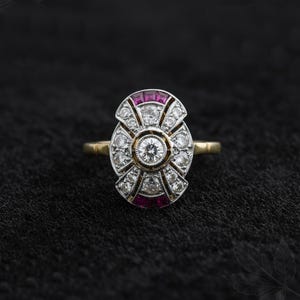 Op de afbeelding: Een antieke ring met een centrale diamant omringd door kleinere diamanten en rode edelstenen. De ring heeft een gouden band en een gedetailleerde, ovale zetting. Het ontwerp is geplaatst tegen een donkere achtergrond, die de details benadrukt.