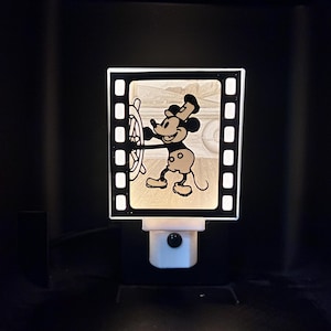 May include: A night light featuring a film strip design with a classic Mickey Mouse image. The black and white image shows Mickey Mouse at the helm of a ship. The night light has a white base and a black border.