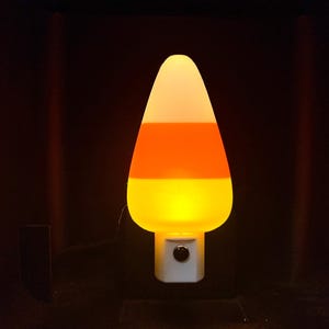 May include: A candy corn-shaped night light with a white base and a three-color design. The top is white, the middle is orange, and the bottom is yellow. The night light is plugged into a wall outlet.