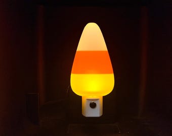 Pode incluir: Uma luz noturna em forma de doce de milho com uma base branca e um design tricolor. A parte superior é branca, o meio é laranja e a parte inferior é amarela. A luz noturna está ligada a uma tomada.