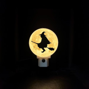 May include: A night light featuring a silhouette of a witch on a broomstick against a full moon. The moon is illuminated, and the witch is black. The night light is white and plugged into an outlet. Halloween decor.