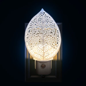 May include: A white, leaf-shaped night light with a detailed, openwork design. The light is plugged into a wall socket and emits a soft, warm glow. The night light is a decorative home accent.