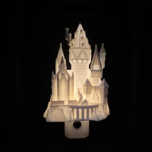 Puede incluir: Una luz de noche con forma de castillo blanco iluminado con intrincados detalles arquitectónicos. El diseño presenta múltiples torres, ventanas arqueadas y un puente, todos iluminados desde dentro sobre un fondo negro. La luz de noche se enchufa a una toma de corriente.