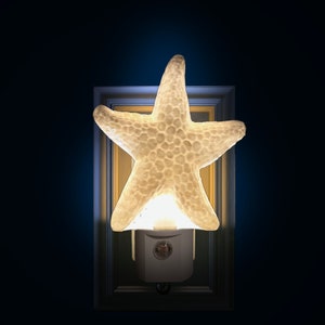 May include: A glowing, white starfish night light. The five-pointed star is textured and sits atop a white base that plugs into a wall outlet. The night light is set against a dark blue background.