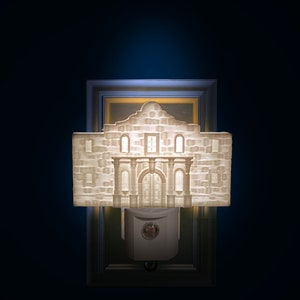 May include: A white, 3D-printed night light shaped like the Alamo, with a detailed facade and arched doorway. The night light is mounted on a white base and is set against a dark blue background. The night light is approximately 5 inches wide and 4 inches tall.