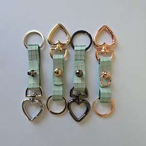 May include: Four keyrings with heart-shaped clasps and decorative hardware. Each keyring has a light green leather strap with a grid pattern. The metal hardware includes silver, gold, black, and rose gold finishes.