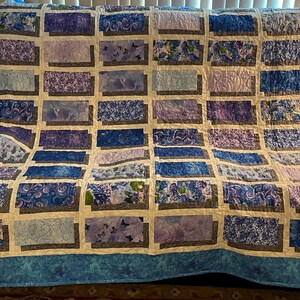 May include: A handmade quilt with a blue and purple color scheme. The quilt features a rectangular pattern with various floral and abstract designs in shades of blue, lavender, and white. The quilt is approximately 80 inches wide and 90 inches long.