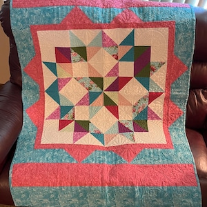 May include: A handmade quilt with a starburst design. The quilt features a central square with geometric patterns in shades of pink, teal, purple, and green. The quilt is bordered by a teal fabric with a pink border and a pink outer edge.