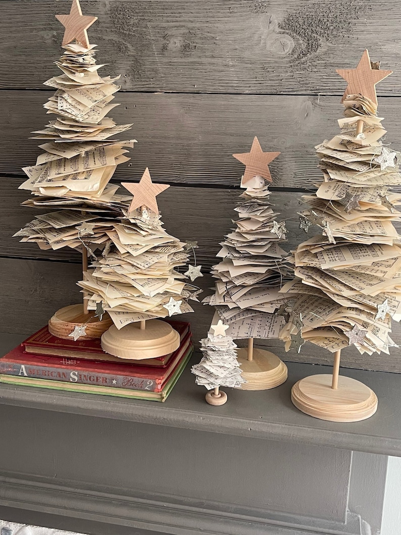May include: Four miniature Christmas trees made of stacked sheet music with wooden bases and star toppers. The trees are arranged on a shelf with a stack of books.