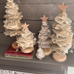 May include: Four miniature Christmas trees made of stacked pages of sheet music with wooden star toppers. The trees are arranged on a gray mantel with a stack of books underneath the largest tree.