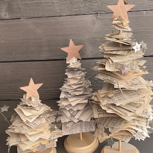 May include: Three miniature Christmas trees made of stacked sheet music with wooden star toppers and a string of silver star lights. The trees are on wooden bases.
