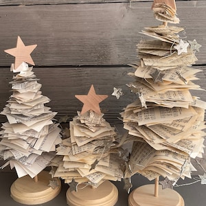 May include: Three miniature Christmas trees made of stacked sheet music with wooden star toppers and silver glitter stars strung around them. The trees are on wooden bases.