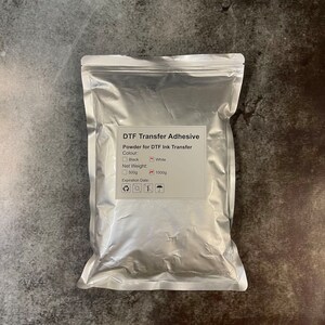 May include: A silver pouch of DTF Transfer Adhesive. The label indicates it's powder for DTF ink transfer, available in black or white. Net weight options are 500g and 1000g. The expiration date is also listed.