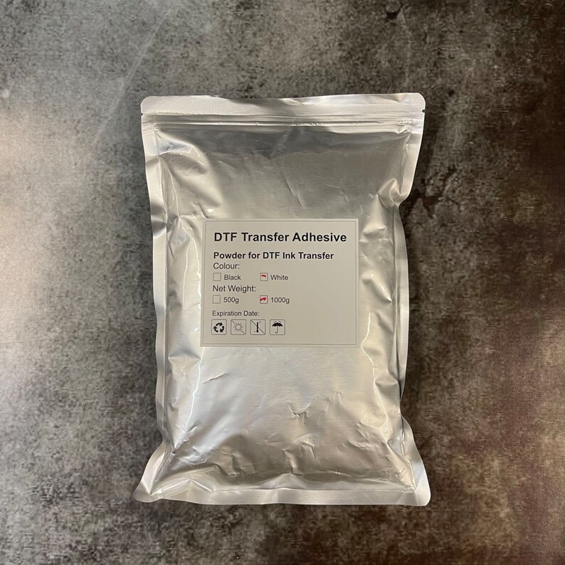 May include: A silver pouch of DTF Transfer Adhesive. The label indicates it's powder for DTF ink transfer, available in black or white. Net weight options are 500g and 1000g. The expiration date is also listed.