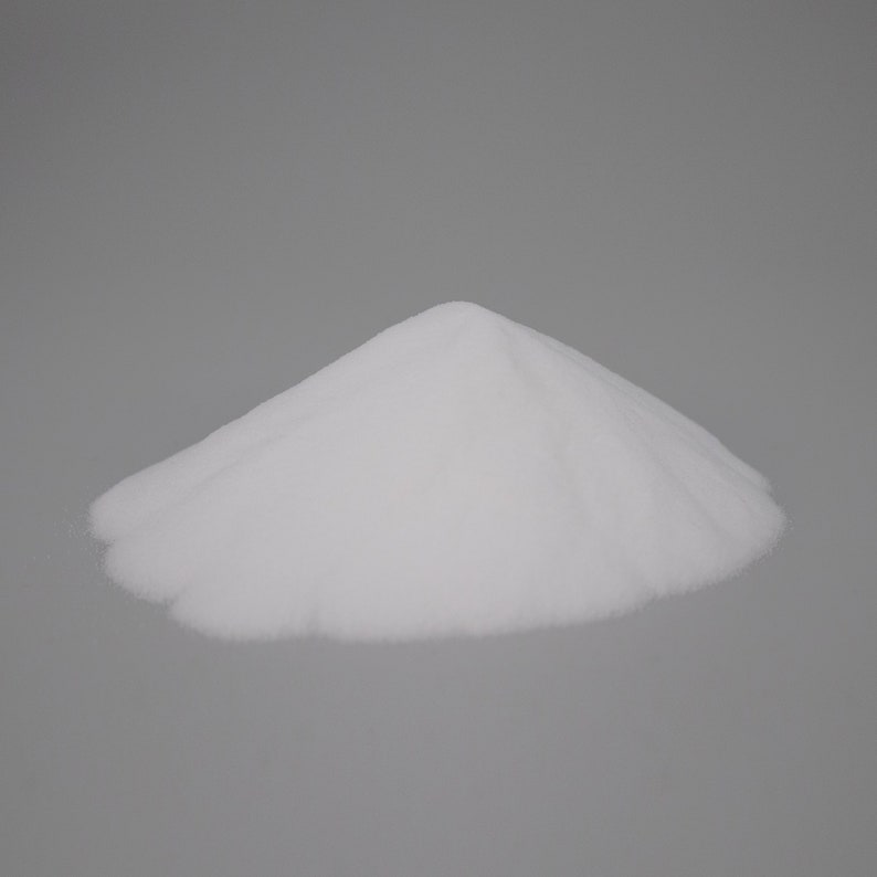 May include: A pile of fine, white powder, possibly a granular substance like sugar or salt, is centered against a plain gray background. The powder forms a small, rounded mountain shape, with soft, textured edges.