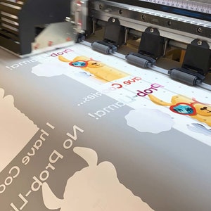 May include: A large format printer printing a design featuring a cartoon llama wearing sunglasses. The design includes the text "I have Cookies... No prob-llama!" in white, along with cloud shapes. The printer is in operation.