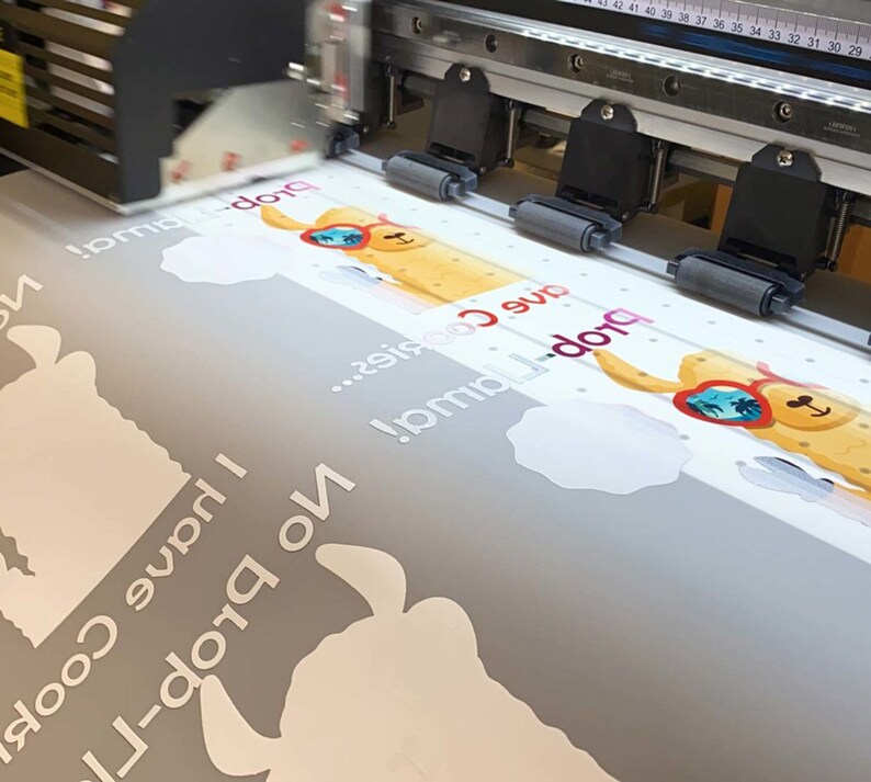 May include: A large format printer printing a design featuring a cartoon llama wearing sunglasses. The design includes the text "I have Cookies... No prob-llama!" in white, along with cloud shapes. The printer is in operation.