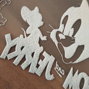 May include: Close-up of a white glittery design featuring the cartoon characters Jerry Mouse and Tom Cat. The word "Jerry" is visible, along with the word "Tom". The design is on a brown background.