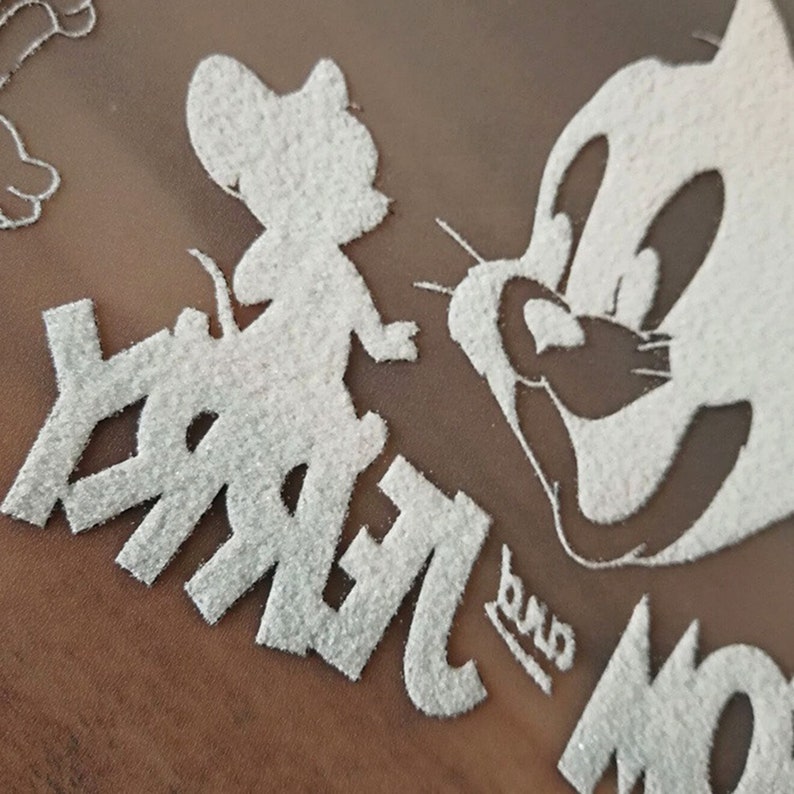 May include: Close-up of a white glittery design featuring the cartoon characters Jerry Mouse and Tom Cat. The word "Jerry" is visible, along with the word "Tom". The design is on a brown background.