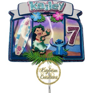 May include: A blue and pink cake topper featuring Lilo and Stitch from Disney's Lilo & Stitch. The topper includes a surfboard, flowers, and the name "Kailey" with the number "7" in pink glitter. The topper is made by Kaybean Creations.