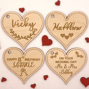K&ouml;nnte beinhalten: Vier herzf&ouml;rmige Holzetiketten mit verschiedenen Botschaften. Das erste Etikett sagt "Vicky" mit einem floralen Design. Das zweite Etikett sagt "Matthew". Das dritte Etikett sagt "Happy 18th Birthday Sophie" mit einem Champagnerglas-Design. Das vierte Etikett sagt "On your wedding day Mr & Mrs Bibbey".