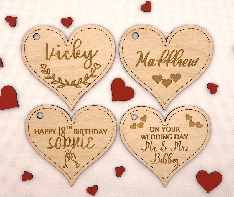 K&ouml;nnte beinhalten: Vier herzf&ouml;rmige Holzetiketten mit verschiedenen Botschaften. Das erste Etikett sagt "Vicky" mit einem floralen Design. Das zweite Etikett sagt "Matthew". Das dritte Etikett sagt "Happy 18th Birthday Sophie" mit einem Champagnerglas-Design. Das vierte Etikett sagt "On your wedding day Mr & Mrs Bibbey".