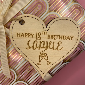 K&ouml;nnte beinhalten: Ein h&ouml;lzerner herzf&ouml;rmiger Geschenkanh&auml;nger mit dem Text "Happy 18th Birthday Sophie" und einer kleinen Illustration von zwei Champagnergl&auml;sern.