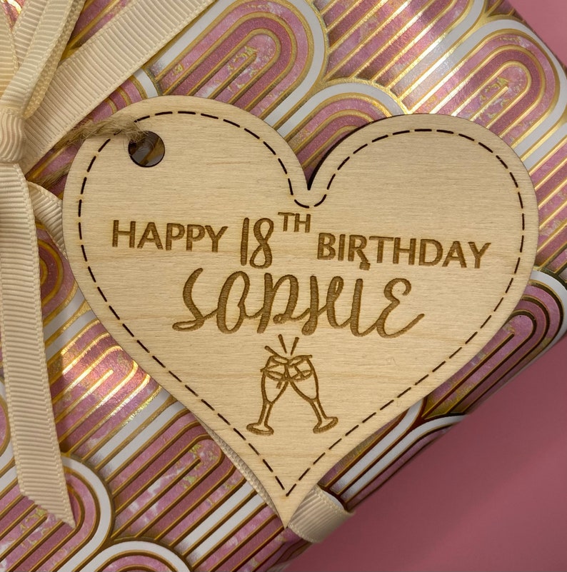 K&ouml;nnte beinhalten: Ein h&ouml;lzerner herzf&ouml;rmiger Geschenkanh&auml;nger mit dem Text "Happy 18th Birthday Sophie" und einer kleinen Illustration von zwei Champagnergl&auml;sern.