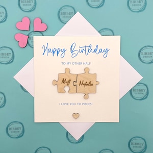 May include: A white card with the text "Happy Birthday" and "To My Other Half" in blue script.  Two wooden puzzle pieces with the names "Matt" and "Natalie" are attached to the card. The card has the text "I Love You To Pieces!" in blue script.