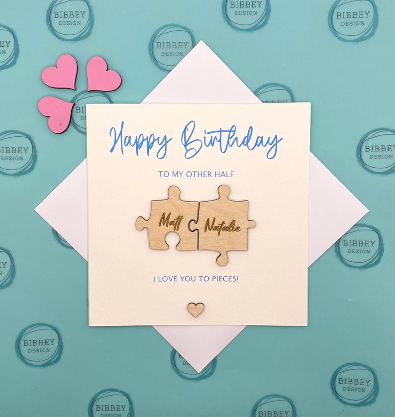 May include: A white card with the text "Happy Birthday" and "To My Other Half" in blue script.  Two wooden puzzle pieces with the names "Matt" and "Natalie" are attached to the card. The card has the text "I Love You To Pieces!" in blue script.