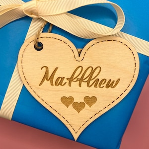 K&ouml;nnte beinhalten: Ein herzf&ouml;rmiges Holzschild mit dem Namen "Matthew" eingraviert. Unter dem Namen sind drei kleine Herzen eingraviert. Das Schild ist mit einem wei&szlig;en Band an einer blauen Geschenkbox gebunden.
