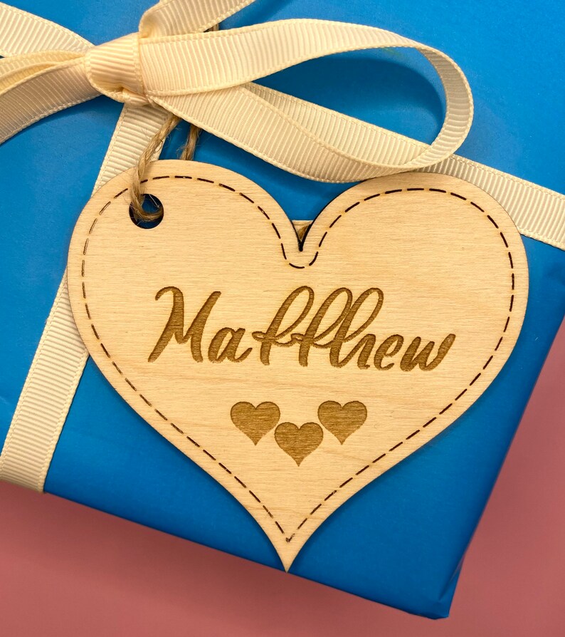 K&ouml;nnte beinhalten: Ein herzf&ouml;rmiges Holzschild mit dem Namen "Matthew" eingraviert. Unter dem Namen sind drei kleine Herzen eingraviert. Das Schild ist mit einem wei&szlig;en Band an einer blauen Geschenkbox gebunden.