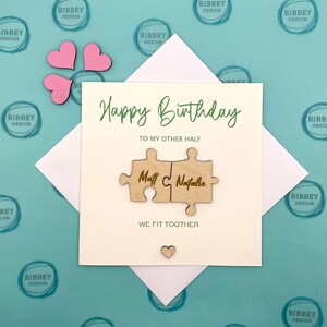 May include: A white card with the text "Happy Birthday" and "To my other half" in green script.  The card features two wooden puzzle pieces that fit together, with the names "Matt" and "Natalie" engraved on them. The text "We fit together" is printed below the puzzle pieces.