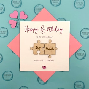 May include: A white card with the text "Happy Birthday" and "To my other half" in pink script. Two wooden puzzle pieces with the names "Matt" and "Natalie" are attached to the card. The card is on a pink and blue background with the words "Bibbey Design" repeated.