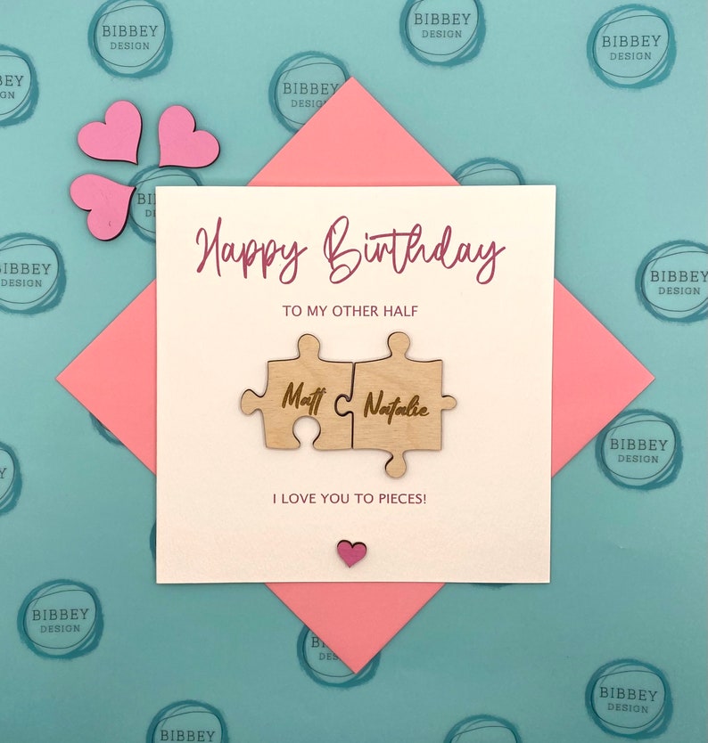 May include: A white card with the text "Happy Birthday" and "To my other half" in pink script. Two wooden puzzle pieces with the names "Matt" and "Natalie" are attached to the card. The card is on a pink and blue background with the words "Bibbey Design" repeated.