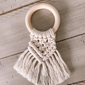 Pu&ograve; includere: Un mordedor per neonati in crochet bianco con un anello di legno e frange. Il mordedor &egrave; realizzato con un motivo a macram&eacute;.