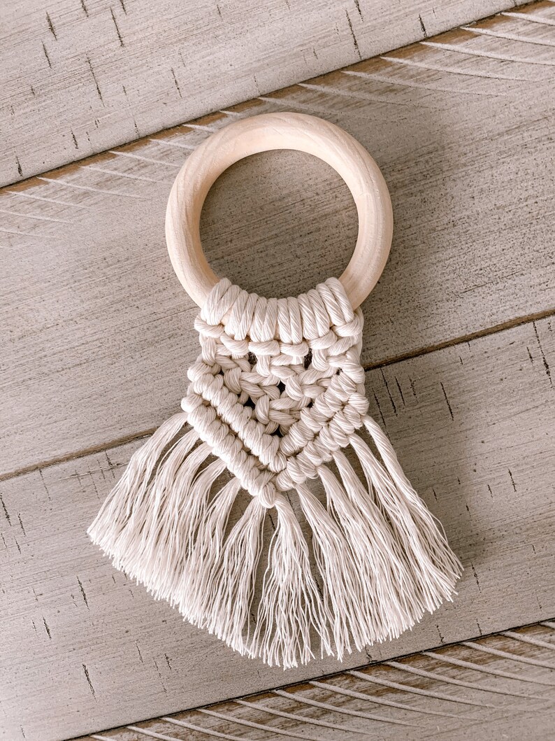 Pu&ograve; includere: Un mordedor per neonati in crochet bianco con un anello di legno e frange. Il mordedor &egrave; realizzato con un motivo a macram&eacute;.
