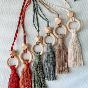 May include: Six wooden ring and bead toys with tassels in various colors, including red, orange, green, and beige. The rings are made of natural wood and the tassels are made of soft yarn.