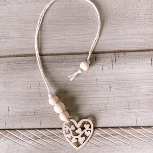 May include: A white wooden heart with a cutout design hangs from a white cord with wooden beads.