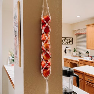 Puede incluir: Una cesta de frutas colgante de macramé blanco con manzanas rojas dentro. La cesta está colgada en una pared de una cocina.