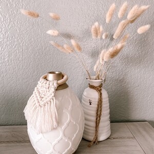 Pu&ograve; includere: Diffusore in ceramica bianca con coperchio dorato e nappa bianca. Il diffusore presenta un motivo decorativo e il testo "Young Living" sul fondo. Il diffusore &egrave; appoggiato su una superficie in legno accanto a un vaso bianco con una corda marrone legata attorno al collo. Il vaso &egrave; pieno di erba della pampa secca.