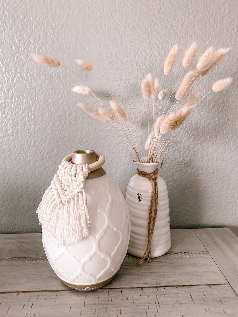 Pu&ograve; includere: Diffusore in ceramica bianca con coperchio dorato e nappa bianca. Il diffusore presenta un motivo decorativo e il testo "Young Living" sul fondo. Il diffusore &egrave; appoggiato su una superficie in legno accanto a un vaso bianco con una corda marrone legata attorno al collo. Il vaso &egrave; pieno di erba della pampa secca.