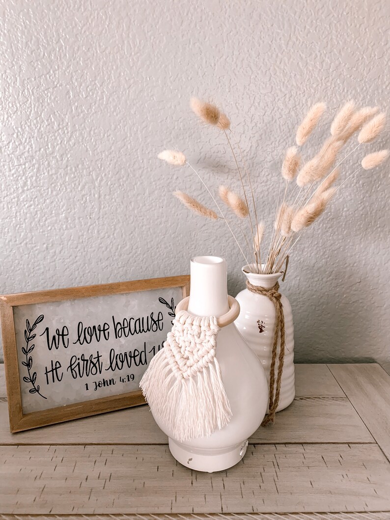 Pu&ograve; includere: Diffusore in ceramica bianca con un anello di legno e un fiocco di macram&egrave; bianco. Il diffusore &egrave; appoggiato su un tavolo di legno con un cartello che recita "We love because He first loved us 1 John 4:19". Dietro il diffusore ci sono erbe della pampa secche in un vaso bianco.
