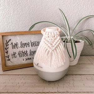 Pu&ograve; includere: Un diffusore bianco con una base grigia e un fiocco di macram&egrave; bianco appeso in cima. Il diffusore &egrave; appoggiato su una superficie di legno con un cartello che recita "We love because He first loved" e una pianta in vaso sullo sfondo.