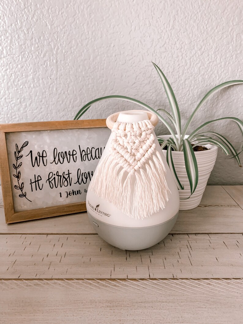 Pu&ograve; includere: Un diffusore bianco con una base grigia e un fiocco di macram&egrave; bianco appeso in cima. Il diffusore &egrave; appoggiato su una superficie di legno con un cartello che recita "We love because He first loved" e una pianta in vaso sullo sfondo.