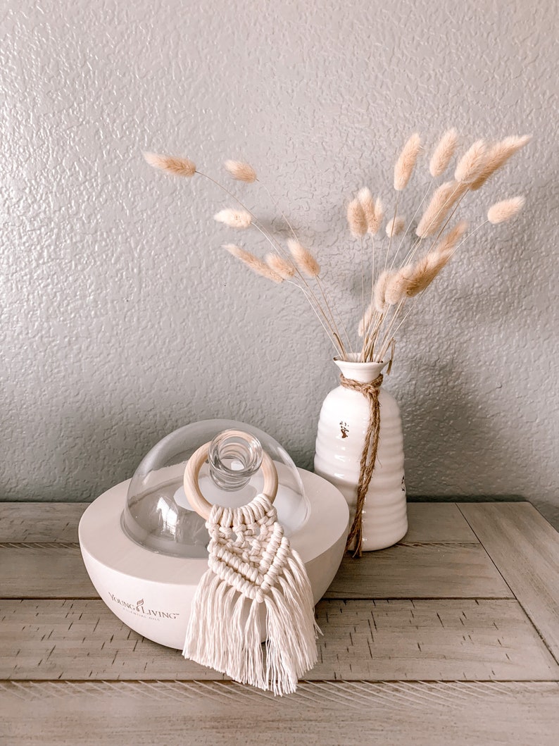 Pu&ograve; includere: Diffusore di oli essenziali in ceramica bianca con una cupola di vetro e un anello di legno. Il diffusore ha la scritta "Young Living" sul davanti. Un fiocco di macram&egrave; bianco pende dal diffusore. Un vaso con erba della pampa secca si trova su un tavolo di legno.