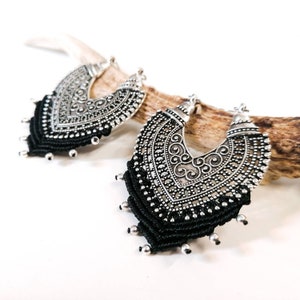 Puede incluir: Un par de pendientes de aro de plata con flecos de macramé negro. Los pendientes tienen un diseño detallado y adornado.